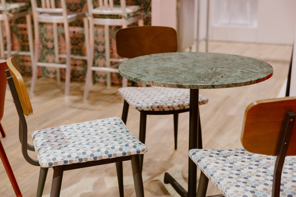 A green marble table top with a black base. 3 chairs sit around the table with wood backrests and a blue and green pattern on the upholstered seat
