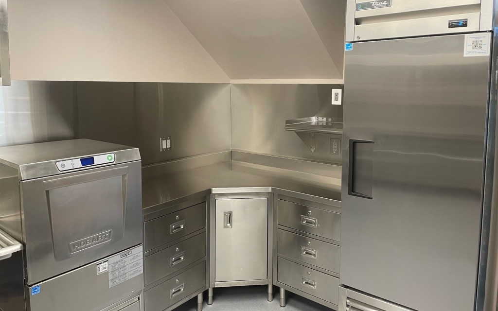 On the left is a stainless steel commercial dishwasher, in the middle there is a custom corner-cabinet lineup with drawers and a cabinet, and immediately to the right is a stainless steel commercial refrigerator.