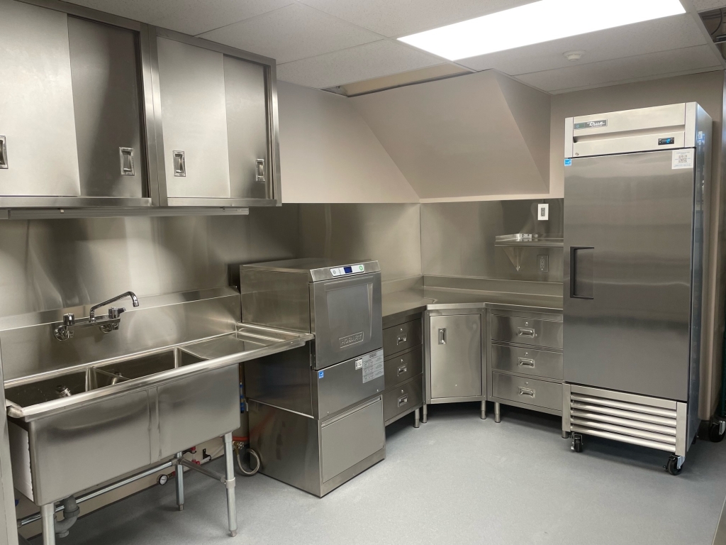 On the left is a stainless steel double compartment sink up against a commercial dishwasher, in the middle there is a custom corner-cabinet lineup with drawers and a cabinet, and immediately to the right is a stainless steel commercial refrigerator.